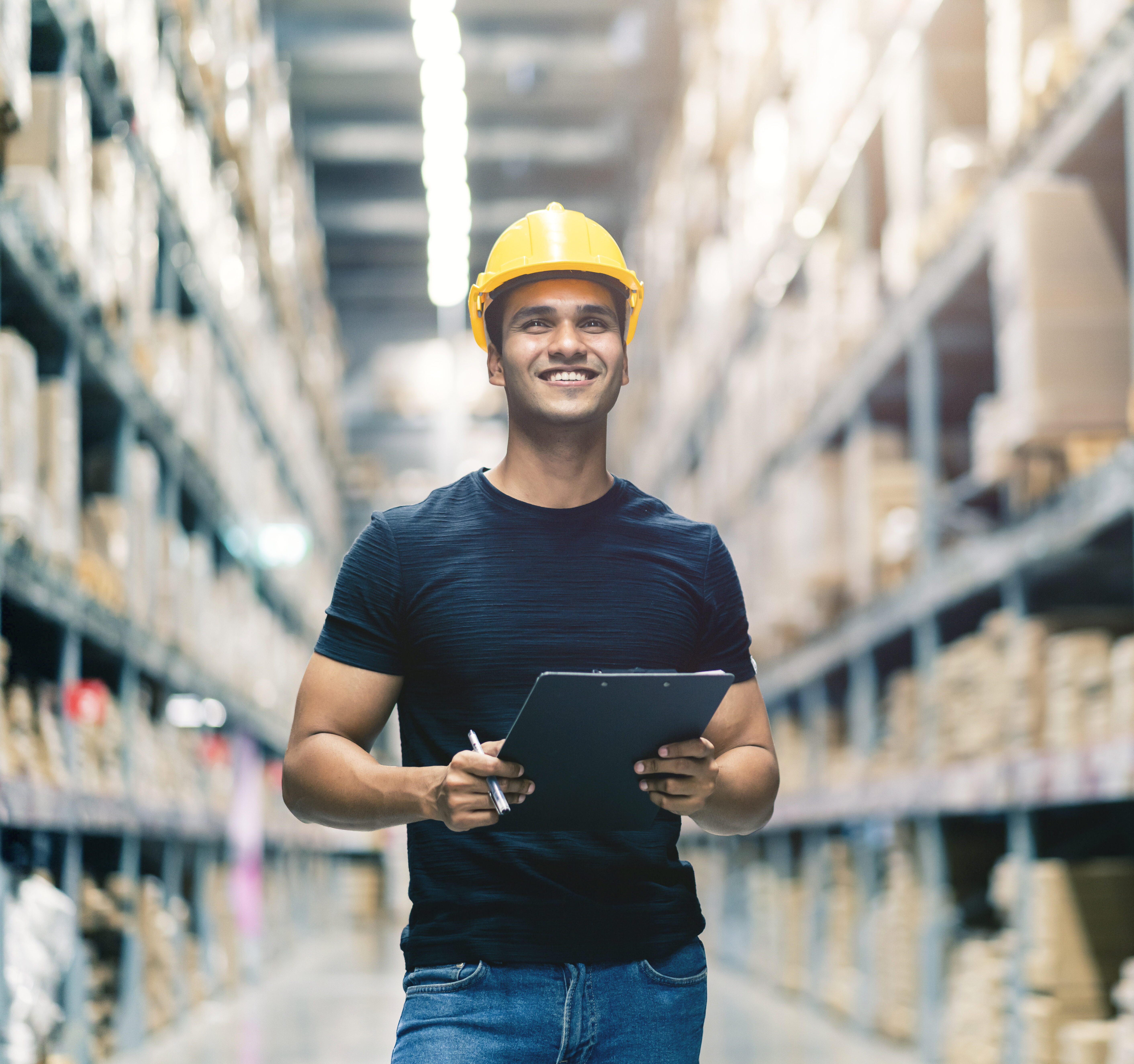 Smart Indian engineer man wearing safety helmet doing stock tick check and cardboard stock product management in factory warehouse background